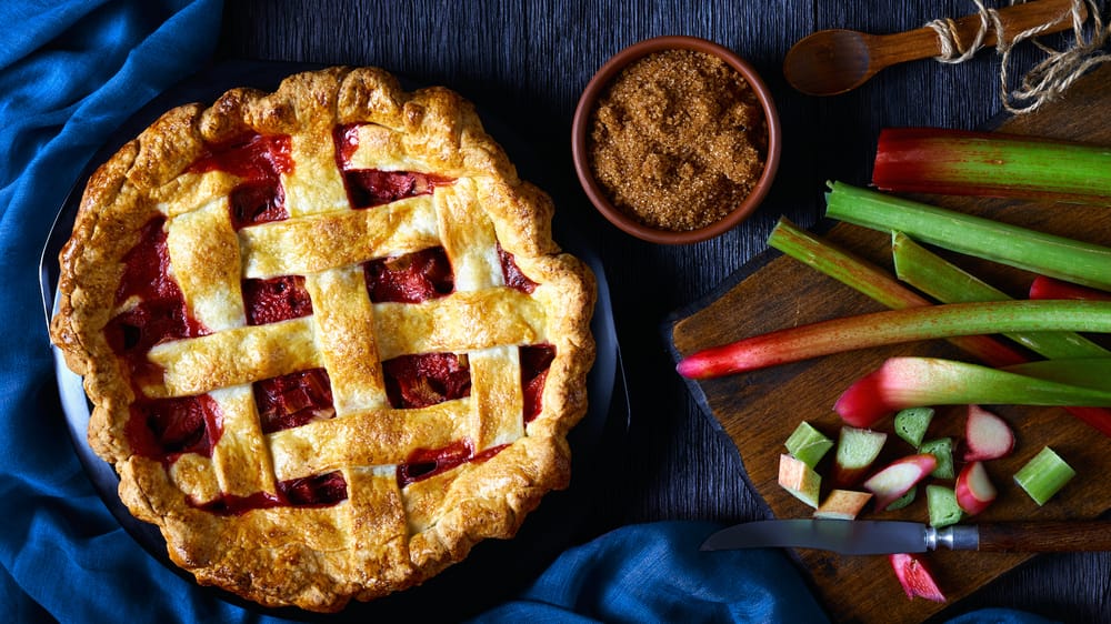 A photograph of a strawberry rhubarb pie on a table, with fresh cut rhubarb and a bowl of cinnamon.